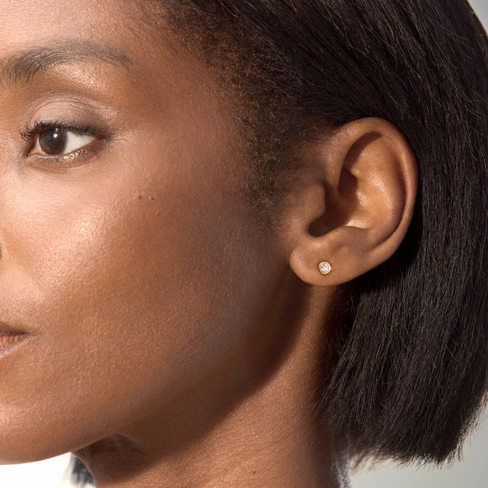 Round bezel-set diamond stud earrings on ear, 18kt gold over sterling finish.