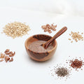 Wooden bowl with a wooden spoon containing a brown spice mix, surrounded by various seeds and spices on a white background.